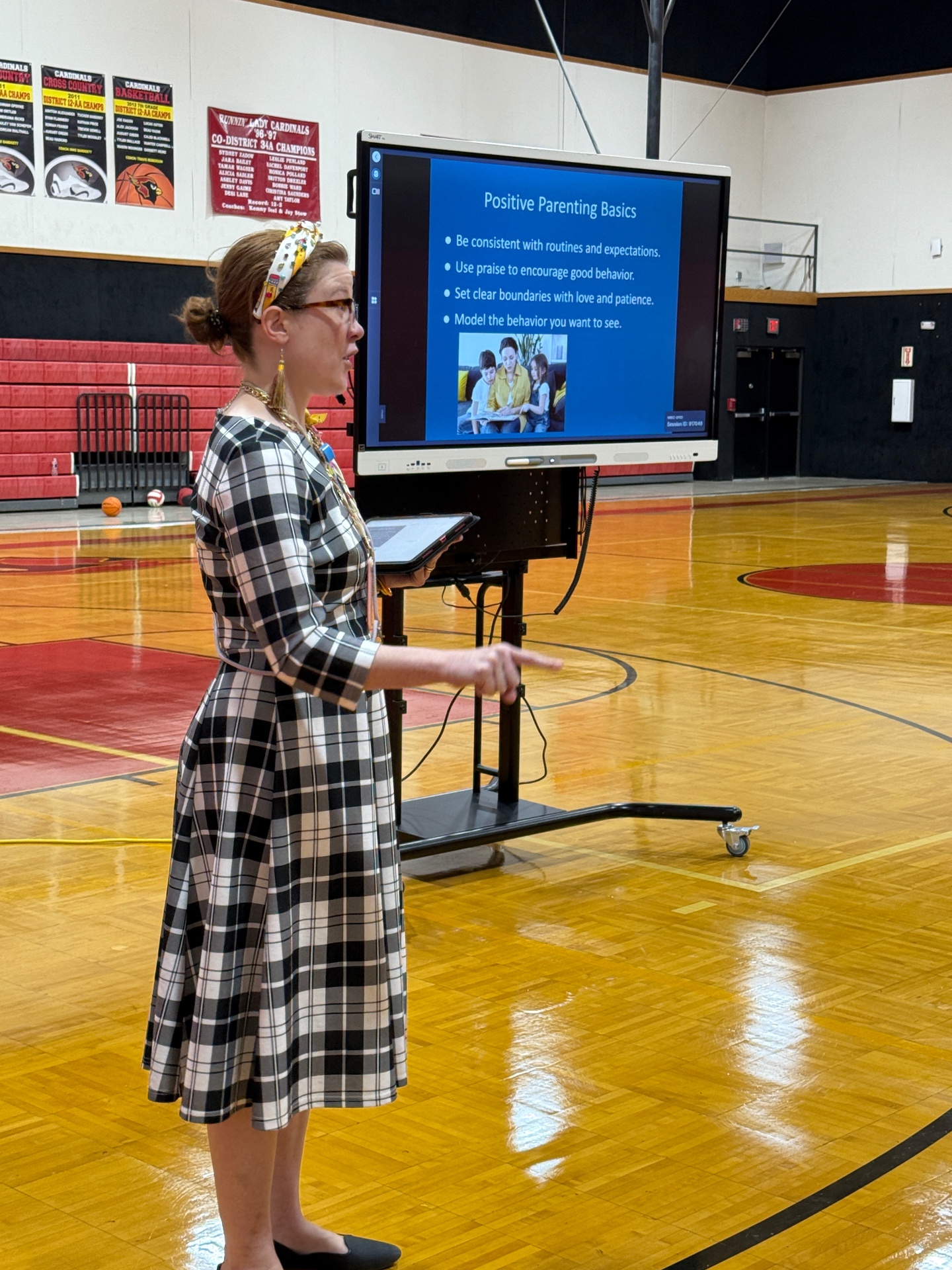 Sally wearing a black and white plaid dress speaking to bilingual pre-K parents in a gym about healthy parenting habits