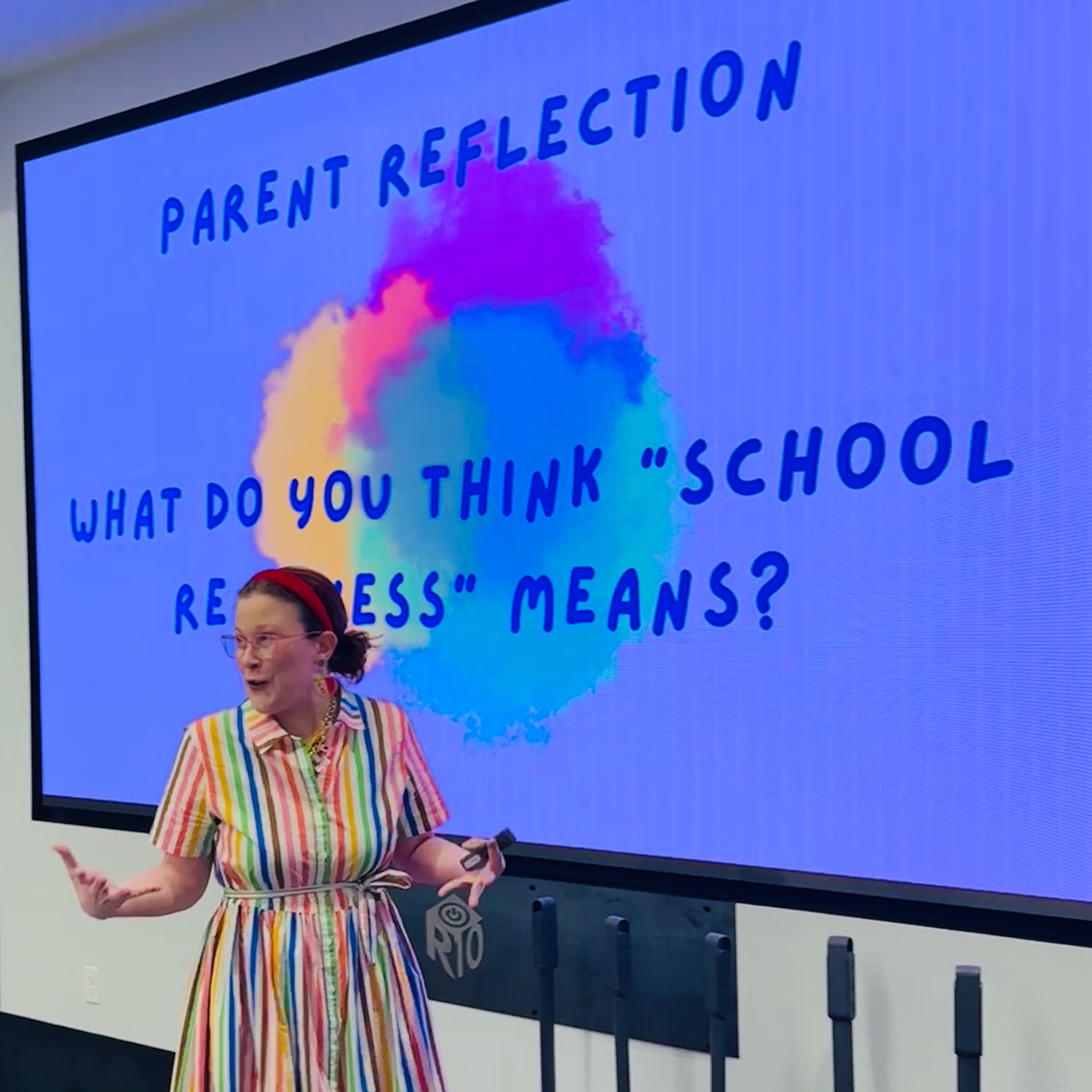 Sally in a vibrant rainbow striped dress speaking about pre-K readiness to parents and teachers at a Title III engagement fair